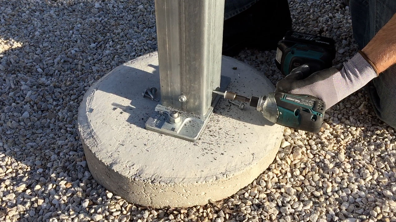 A man attaches an SHS steel post to an Ezipier pier base with screws, on a concrete pad footing using a cordless electric drill.